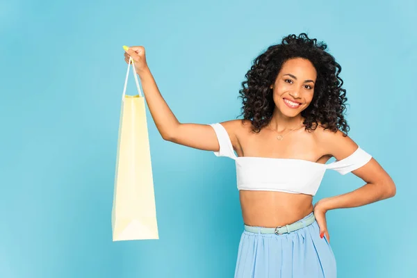 Fröhliches afrikanisch-amerikanisches Mädchen, das mit der Hand auf der Hüfte steht und die Einkaufstasche isoliert auf blau hält — Stock Photo