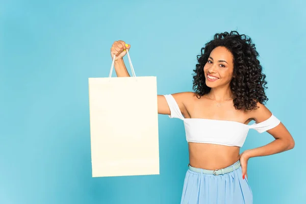 Feliz afroamericana chica de pie con la mano en la cadera y la celebración de bolsa de compras aislado en azul - foto de stock