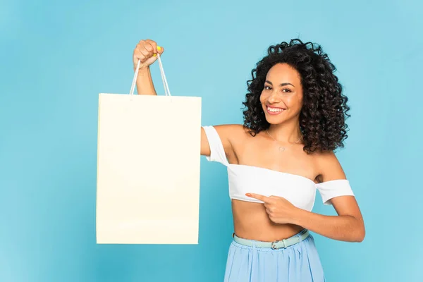 Feliz afroamericana chica apuntando con el dedo a la bolsa de compras aislado en azul - foto de stock