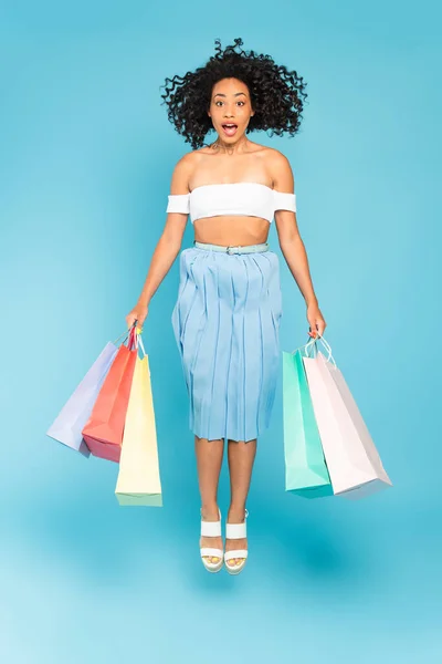 Menina americana africana surpreso segurando sacos de compras e pulando em azul — Fotografia de Stock