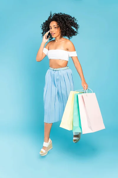 Encaracolado menina americana africana segurando sacos de compras e falando no smartphone em azul — Fotografia de Stock