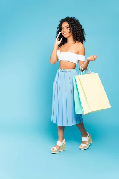 Chica afroamericana positiva sosteniendo bolsas de compras y hablando en el teléfono inteligente en azul - foto de stock