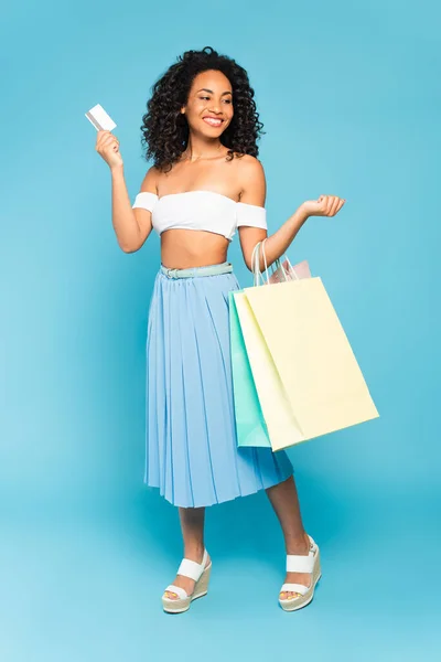 Chica afroamericana positiva sosteniendo bolsas de compras y tarjeta de crédito en azul - foto de stock