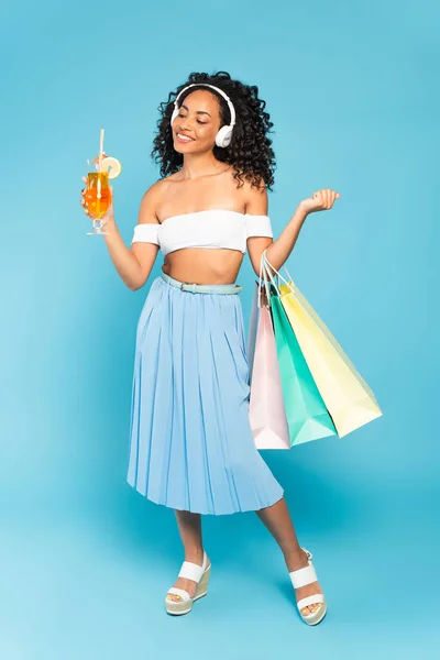 Happy african american girl holding shopping bags and cocktail while listening music in headphones on blue — Stock Photo