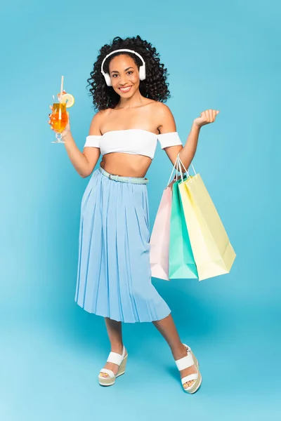 Alegre afroamericana chica sosteniendo bolsas de compras y cóctel mientras escucha música en auriculares en azul - foto de stock