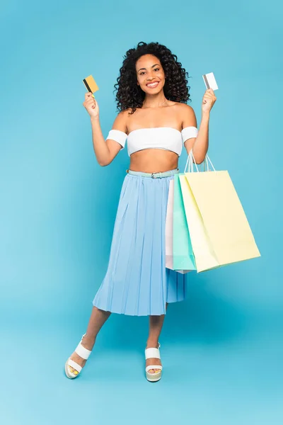 Menina americana africana feliz segurando cartões de crédito e sacos de compras enquanto está em pé no azul — Fotografia de Stock