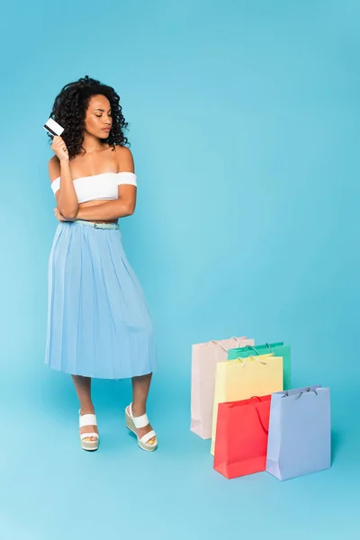 Afrikanerin hält Kreditkarte in der Hand und betrachtet Einkaufstüten, während sie auf blauem Grund steht — Stock Photo