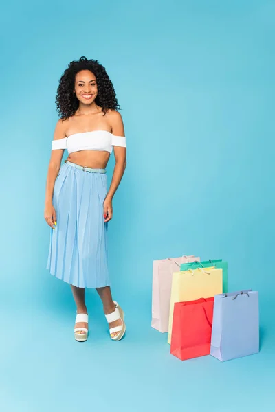 Feliz afroamericana chica de pie cerca de bolsas de compras en azul — Stock Photo
