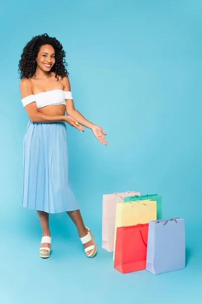 Glückliches afrikanisch-amerikanisches Mädchen stehend und mit den Händen auf Einkaufstüten auf blau zeigend — Stockfoto