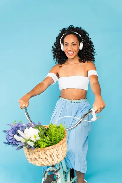 Alegre afroamericana chica en auriculares a caballo bicicleta en azul, concepto de verano - foto de stock