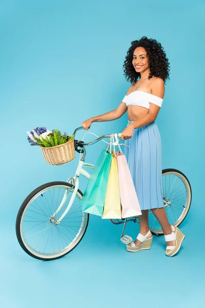 Cheerful african american girl standing near bicycle with shopping bags on blue, summer concept — Stock Photo