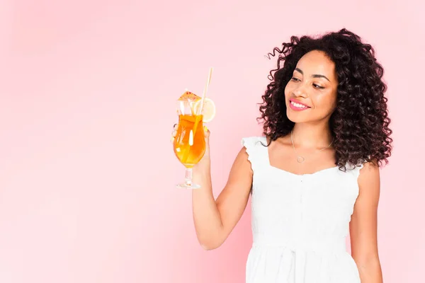 Весёлая африканская американка смотрит на стакан с коктейлем на розовом — стоковое фото