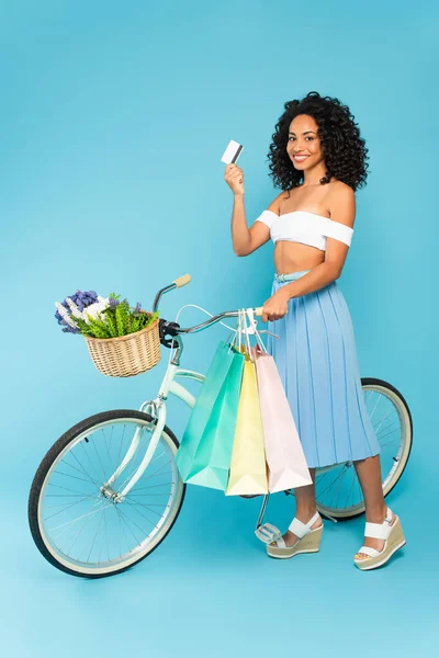 Menina americana africana alegre de pé perto de bicicleta e segurando cartão de crédito e sacos de compras em azul, conceito de verão — Fotografia de Stock