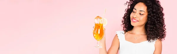 Concetto panoramico di allegra ragazza afroamericana guardando vetro con cocktail isolato su rosa — Foto stock