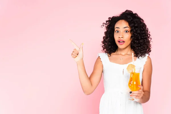 Surprised african american girl pointing with finger and holding cocktail on pink — Stock Photo