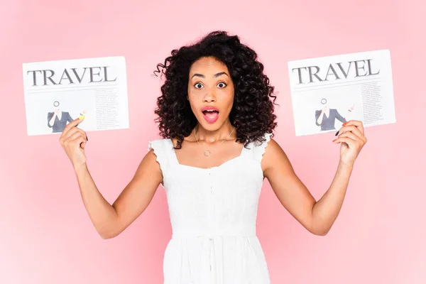 Mujer afroamericana conmocionada sosteniendo periódicos de viaje aislados en rosa - foto de stock