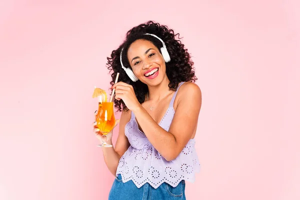 Cheerful african american girl in wireless headphones listening music and holding cocktail isolated on pink — Stock Photo