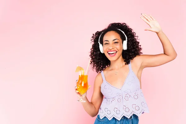 Cheerful african american girl in wireless headphones listening music, gesturing and holding cocktail isolated on pink — Stock Photo