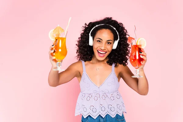 Fröhliche afrikanisch-amerikanische Mädchen in drahtlosen Kopfhörern Musik hören und halten Cocktails isoliert auf rosa — Stockfoto