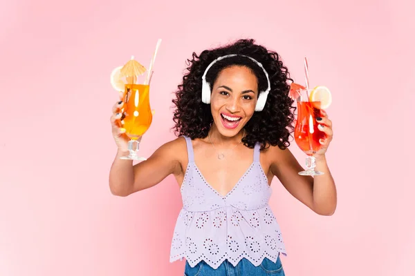 Menina americana africana alegre em fones de ouvido sem fio ouvir música e segurando coquetéis isolados em rosa — Fotografia de Stock