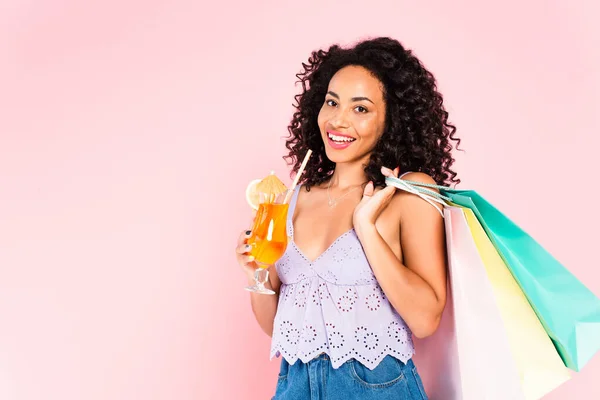 Chica afroamericana feliz sosteniendo bolsas de compras y cóctel aislado en rosa - foto de stock