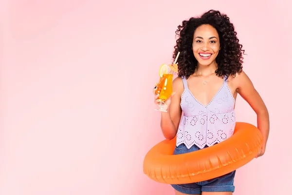 Sorrindo mulher afro-americana de pé com anel de natação e segurando coquetel isolado em rosa — Fotografia de Stock