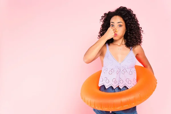 Mujer afroamericana de pie con anillo de natación y la nariz tocando aislado en rosa - foto de stock