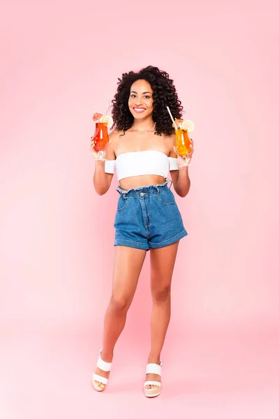 Lächelnde afrikanisch-amerikanische Frau steht und hält Cocktails auf rosa — Stockfoto