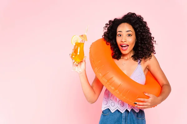 Excited african american woman standing with swim ring and holding cocktail isolated on pink — Stock Photo