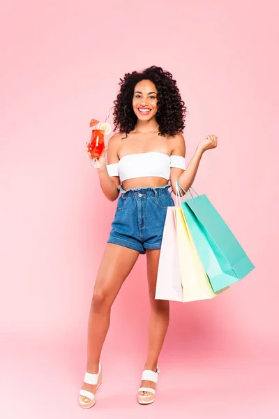 Mujer afroamericana feliz celebración de cócteles y bolsas de compras en rosa - foto de stock
