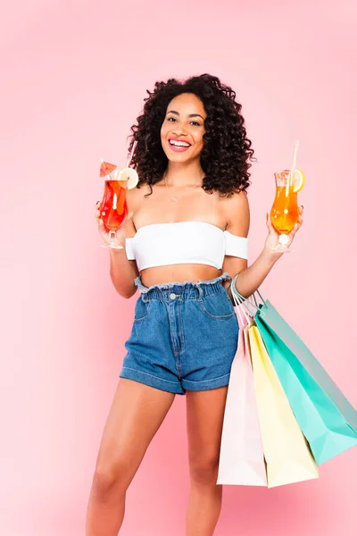 Happy african american woman holding cocktails and shopping bags on pink — Stock Photo