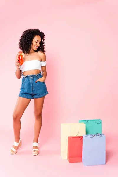 Mujer afroamericana feliz sosteniendo cóctel y de pie con la mano en el bolsillo mientras mira las bolsas de compras en rosa - foto de stock