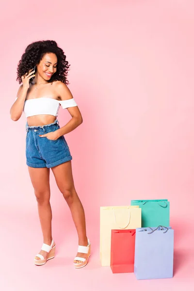 Cheerful african american woman talking on smartphone and standing with hand in pocket near shopping bags on pink — Stock Photo