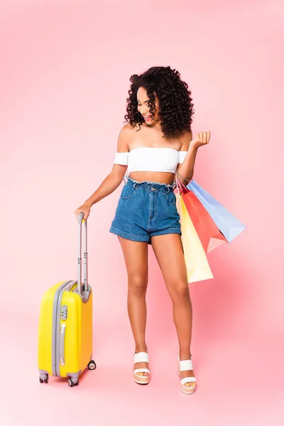 Happy  african american woman standing with luggage and holding shopping bags on pink — Stock Photo