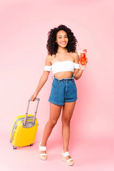 Alegre afroamericana mujer celebración cóctel y caminar con maleta en rosa - foto de stock