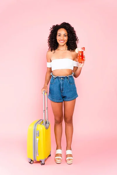 Усміхнена афроамериканка тримає коктейль і стоїть з валізою на рожевому — стокове фото