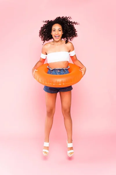 Smiling african american woman holding swim ring while jumping on pink — Stock Photo