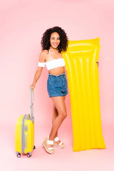 Smiling african american girl holding inflatable mattress and standing near luggage on pink — Stock Photo
