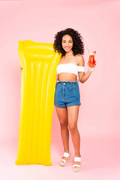 Smiling african american girl holding cocktail and inflatable mattress on pink — Stock Photo