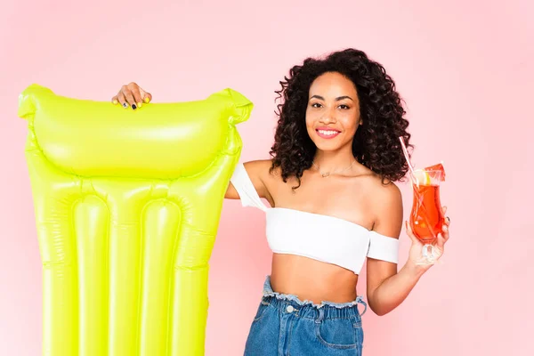 Heureux afro-américaine fille debout près matelas gonflable et tenant cocktail isolé sur rose — Photo de stock