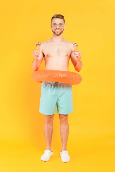 Homem feliz e sem camisa em óculos de sol com anel de natação segurando cones de sorvete no amarelo — Stock Photo