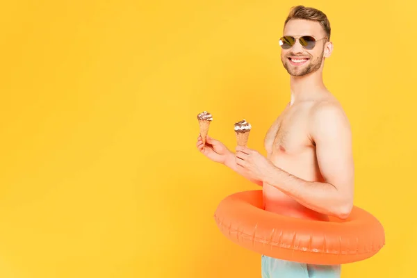 Веселий і без сорочки чоловік в сонцезахисних окулярах з плавальним кільцем, що тримає конуси морозива ізольовані на жовтому — стокове фото