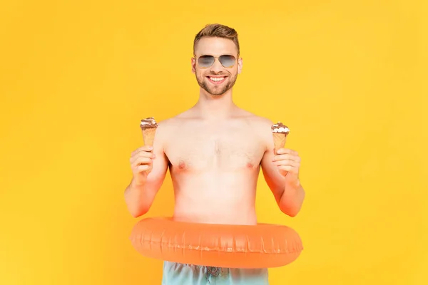 Homem alegre e sem camisa em óculos de sol com anel inflável segurando cones de sorvete isolados no amarelo — Fotografia de Stock