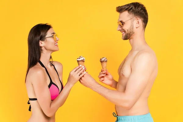 Профиль счастливой девушки, принимающей мороженое рядом с мускулистым мужчиной в солнечных очках, изолированных на желтом — стоковое фото