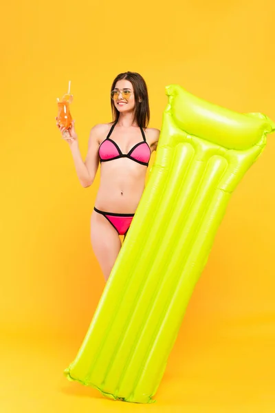 Happy girl in sunglasses looking at cocktail and holding inflatable mattress on yellow — Stock Photo