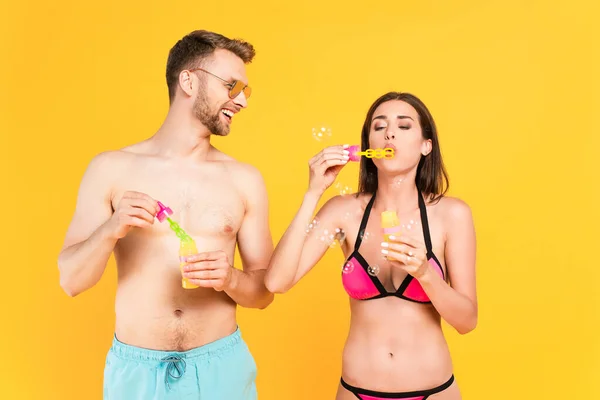 Hombre feliz y sin camisa en gafas de sol mirando a la chica en traje de baño soplando burbujas de jabón aislado en amarillo - foto de stock