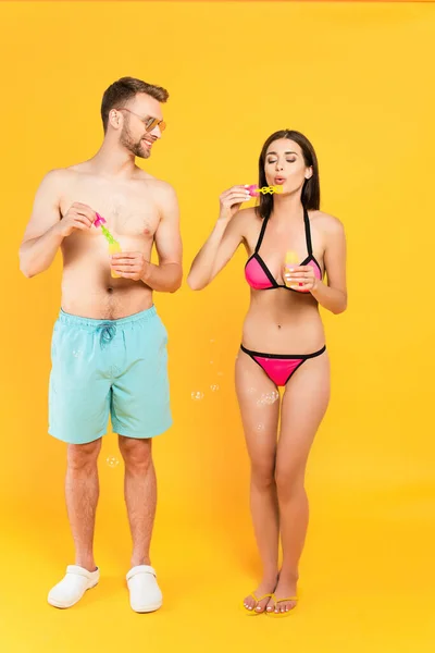 Happy and shirtless man in sunglasses looking at girl in swimsuit blowing soap bubbles on yellow — Stock Photo