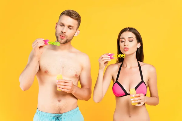 Shirtless man and attractive girl in swimsuit blowing soap bubbles isolated on yellow — Stock Photo