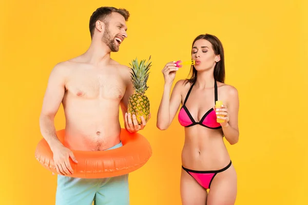 Glücklicher und hemdloser Mann mit Schwimmring, der Ananas in der Nähe eines Mädchens im Badeanzug hält, das Seifenblasen bläst, isoliert auf gelb — Stockfoto