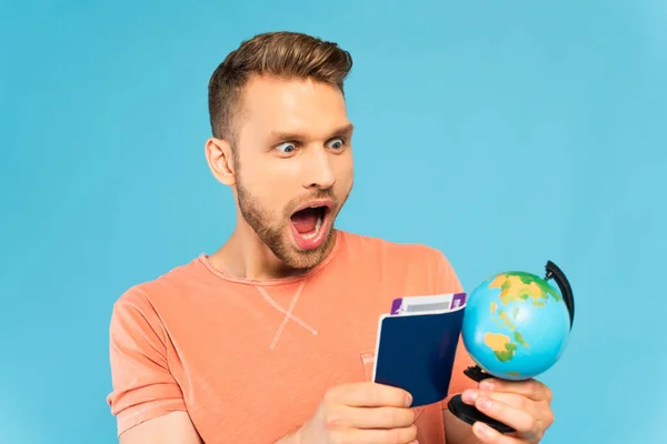 Homem barbudo chocado segurando passaporte e olhando globo isolado em azul — Fotografia de Stock